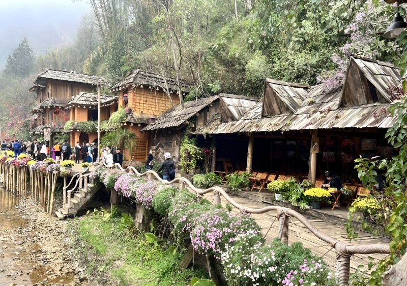 Black Hmong elderly woman in indigo-dyed traditional clothing standing in front of a wooden stilt house at Cat Cat Village 19th century settlement