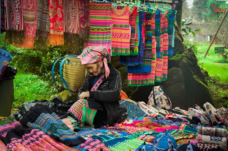 Black Hmong woman demonstrating traditional indigo fabric dyeing at Cat Cat Village cultural experience station with hand-loom weaving in background