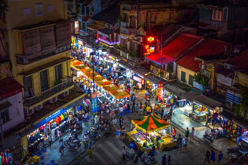 Hanoi Night Market vendor stalls on Hàng Đào Street at night with áo dài clothing, silk scarves, lacquerware, and crowds of weekend shoppers