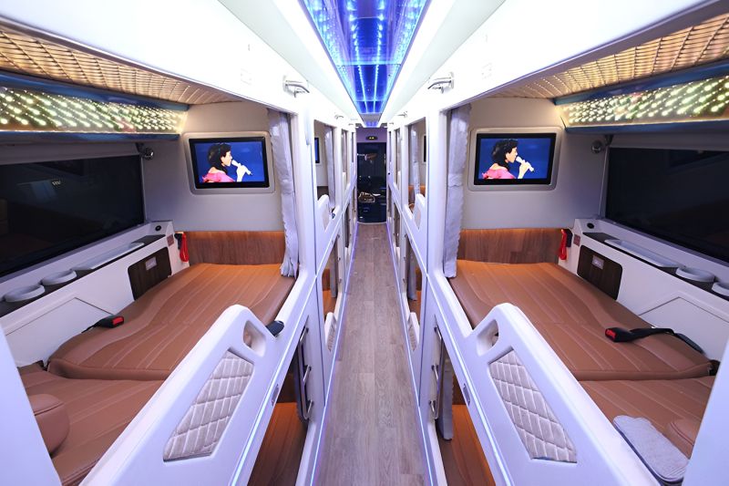 Interior of a premium sleeper bus showing two rows of brown leather semi-sleeper berths with individual TV screens and blue LED ceiling lighting
