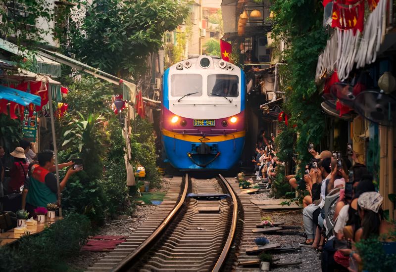 Hanoi Train Street daytime train passing through narrow Phung Hung residential lane with visitors and greenery