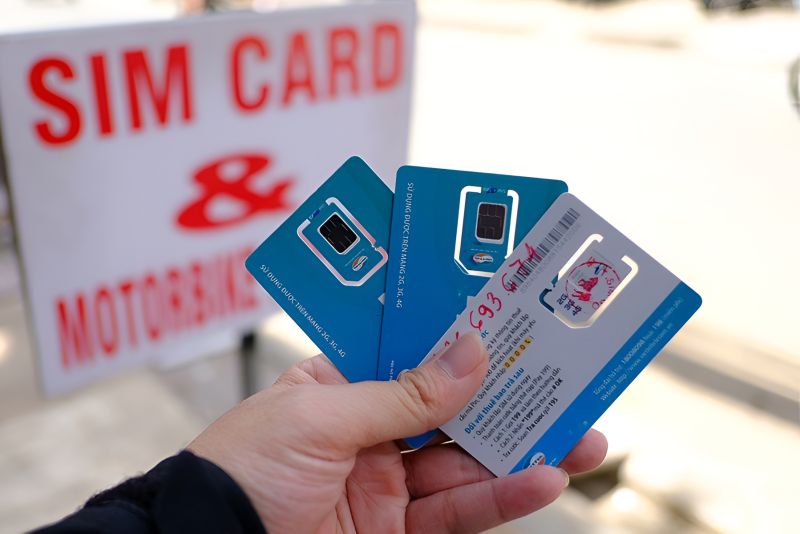 Hand holding three Vietnamese prepaid SIM cards with nano chips visible in front of a street vendor sign advertising SIM cards