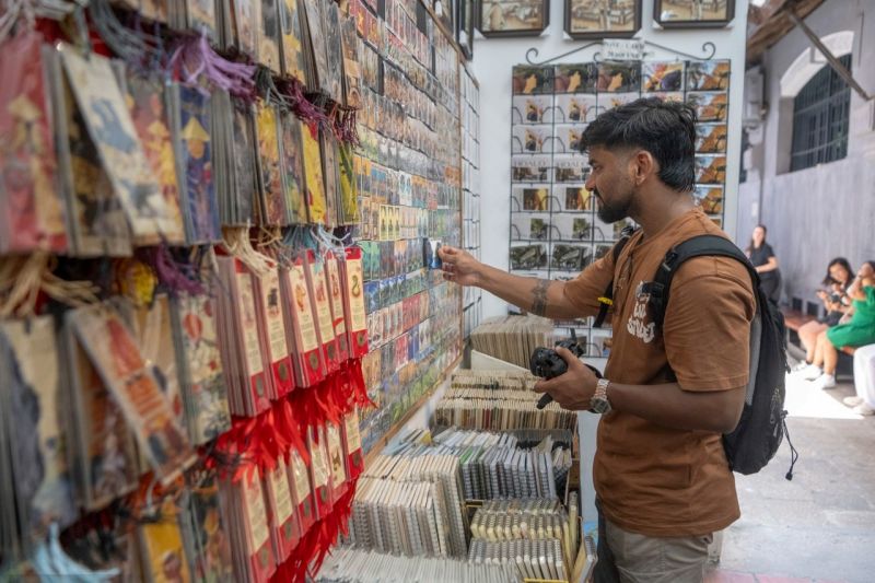 Male tourist with backpack and camera examining postcards and souvenir items on wall display at Vietnamese specialty shop