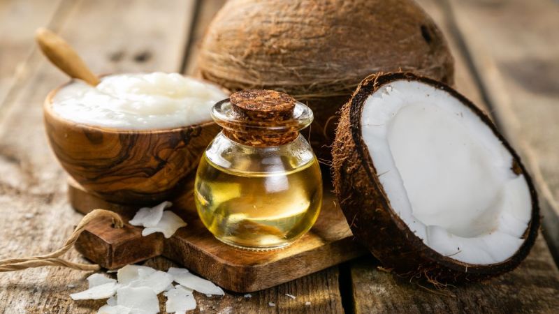 Glass bottle of cold pressed coconut oil on wooden cutting board beside halved fresh coconut and coconut cream in wooden bowl