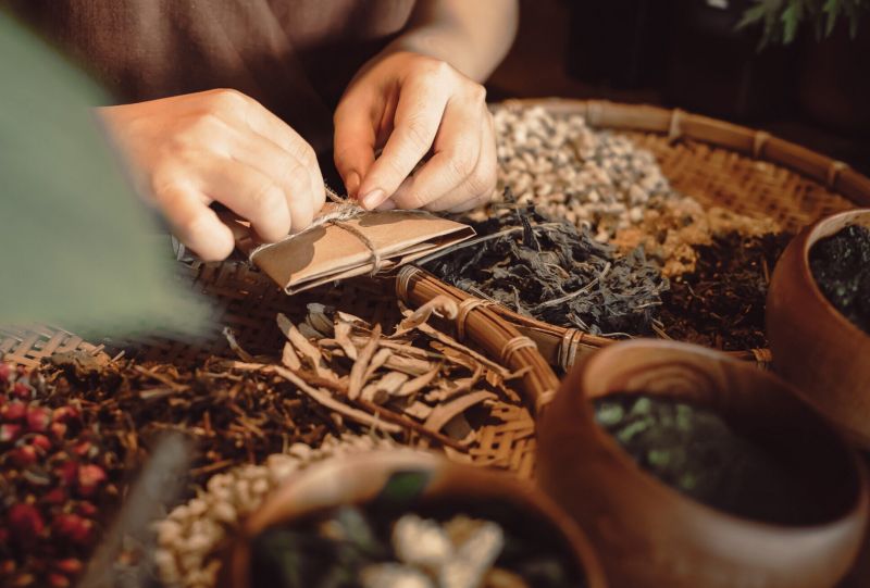 Hands wrapping dried herbs above bamboo tray containing loose tea leaves dried bark red berries and clay pots of herbal ingredients