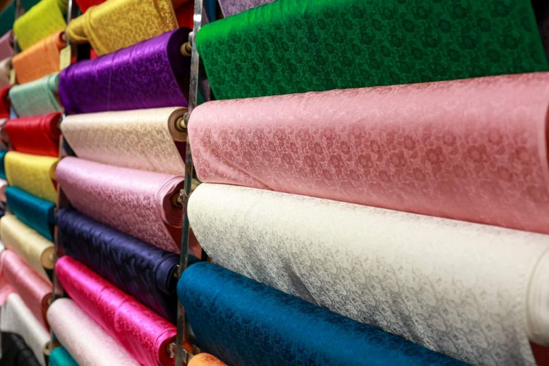 Rows of silk and brocade fabric rolls in pink green blue gold and white with floral embossed patterns on metal display rack