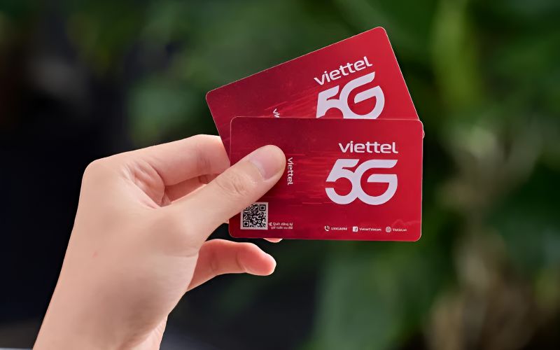 Hand holding two red Viettel 5G prepaid SIM cards with QR code and Viettel branding against green foliage background
